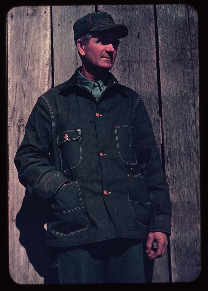 40s Fashion Men Bill Cap Dungaree Coat Wearing attire that would suggest he's a farmer, a man wears a bill cap and a dungaree coat over a chambray shirt circa 1941-42.