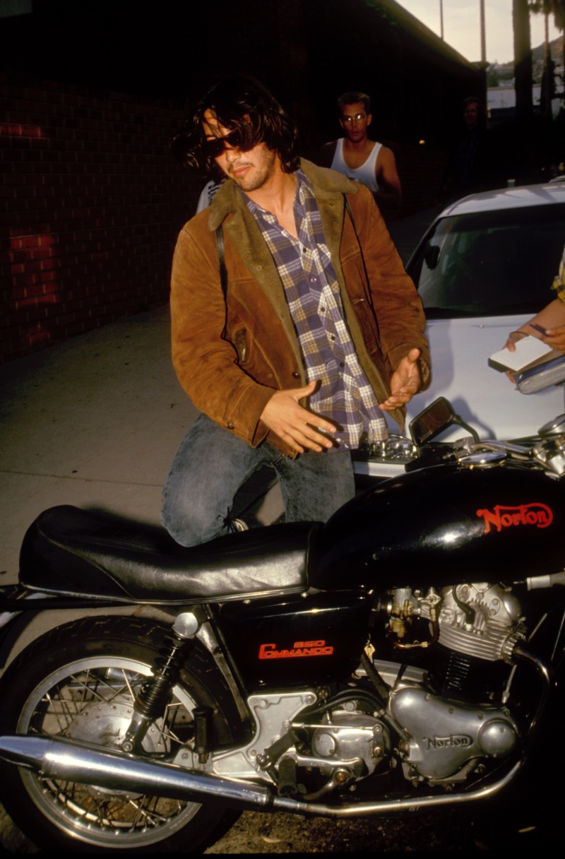 Keanu Reeves 1990s Mens Fashion Shearling Jacket Pictured circa 1991, Keanu Reeves wears a shearling jacket with a plaid button-down as he gets on his motorcycle.